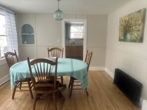 A dining area with a round table covered by a light blue tablecloth, surrounded by wooden chairs. There is a pendant light above the table, a radiator on the wall, and a painting above the radiator. - Sawyer Cove Cottage (Jonesport)