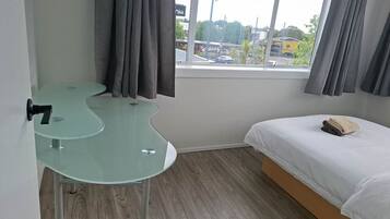 A hotel room with a curved glass table, a bed with a white mattress and a beige blanket, and a window with curtains.