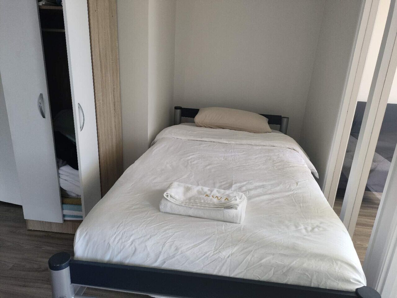 Una cama bien hecha con una toalla doblada, un armario con espejo, y una puerta que da a otra habitación.