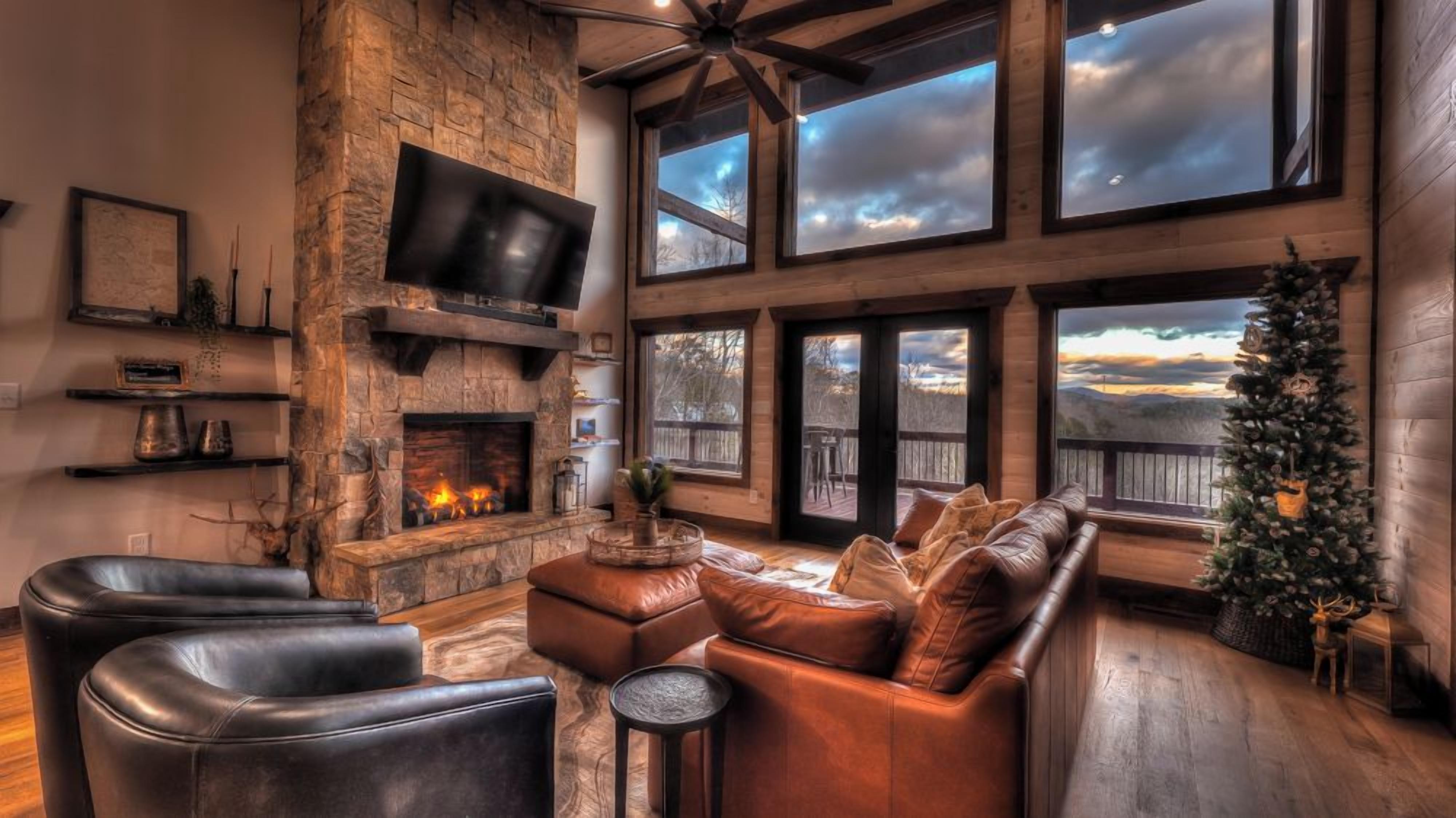 A cozy living room with leather furniture, a fireplace, and large windows offering a view of the outdoors.
