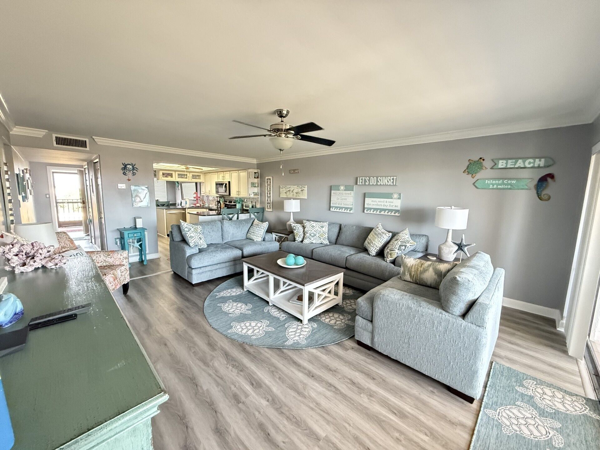 A living room with a grey sectional sofa, a coffee table, and a patterned rug. There is a ceiling fan, a kitchen in the background, and a wall with a 'BEACH' sign.