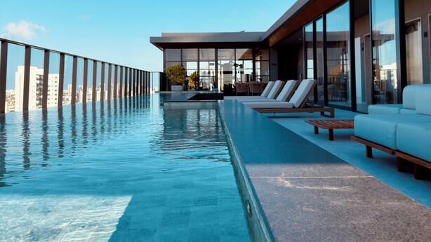 Una moderna piscina a sfioro con vista chiara sullo skyline cittadino, un’area prendisole e un edificio contemporaneo con ampie vetrate.