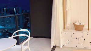 Un balcone con vista sulla città illuminata di notte, una sedia bianca, un tavolo, un appendiabiti e un tappeto tessuto.