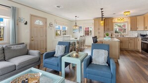 A spacious living room with a grey sofa, two blue armchairs, and a small coffee table. A dining area with a white table and chairs is visible in the background. The kitchen area features wooden cabinets and a large island with a sink. - 5206 Hogg Heaven * 3 Min Walk to Beach (Kill Devil Hills)