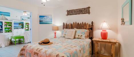 A bedroom with a beach-themed bedspread, a ceiling fan, a wooden headboard, a bedside table with a lamp, and a bookshelf filled with books and decorative items.