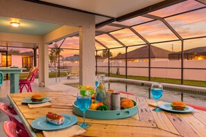 An outdoor dining area with a pool view, tables with food and drinks, and a sunset in the background. - Heated Pool, Lanai, Grill and Games - Villa Sandy Cheeks - Roelens Vacations (Cape Coral)