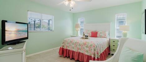 A bedroom with a bed, a television, a ceiling fan, and a window with blinds.