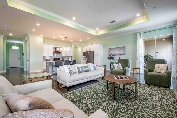 A spacious living room with a sofa, a coffee table, and a patterned rug. There is a kitchen area with cabinets and appliances in the background.