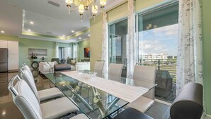 Un área de comedor moderna con una mesa y sillas de vidrio, una araña y vistas de la ciudad a través de grandes ventanales.