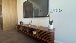 Een woonkamer met een flatscreen televisie aan de muur, een houten console en decoratieve objecten, en een potorkidee.
