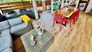 A spacious living area with a wooden floor, a sofa, a dining table with red chairs, and a kitchen area in the background.