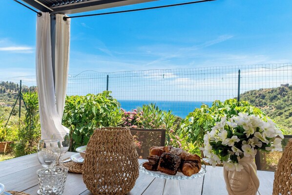 A table with glassware, pastries, and a vase of flowers, overlooking a scenic coastal landscape.