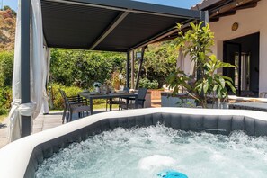 Ein Whirlpool mit Tisch und Stühlen unter einer überdachten Terrasse, umgeben von Grünpflanzen.