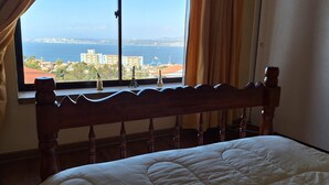 A bedroom with a wooden headboard, a bed with a white quilt, and a view of the sea and buildings through a window with curtains.