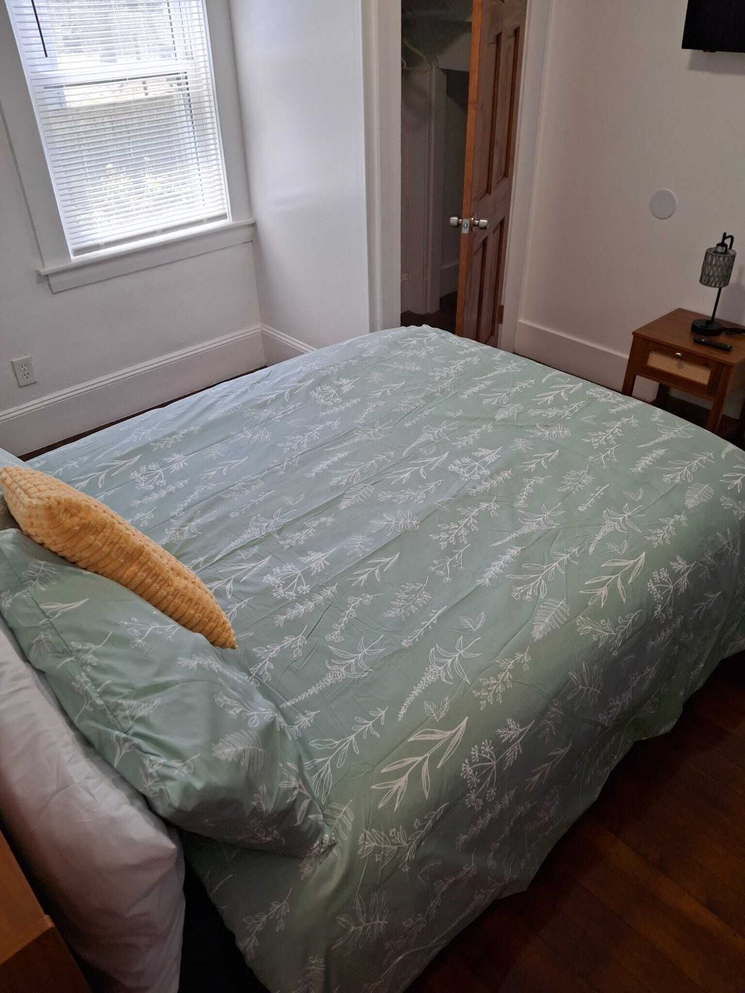 Une pièce avec un lit, une fenêtre, une porte et une petite table avec une lampe.