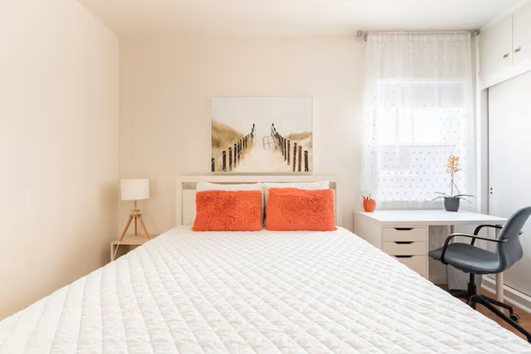 Ein Schlafzimmer mit einem Bett, zwei orangefarbenen Kissen, einem Schreibtisch mit Stuhl und einem Fenster mit Vorhängen.