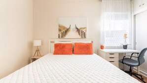Une chambre à coucher avec un lit, deux oreillers orange, un bureau avec une chaise et une fenêtre avec des rideaux.
