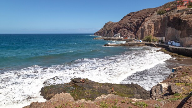 A coastal landscape with rocky terrain, a clear sky, and a building with a red roof near the water.