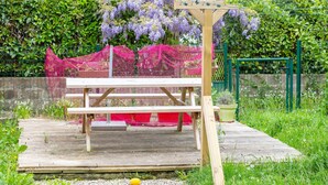 A wooden bench under a pergola with purple flowers, surrounded by a garden with a fence and greenery.