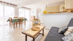 Un salon moderne comprenant un canapé, une table à manger et des chaises, un coin cuisine avec des meubles en bois, et une grande fenêtre avec des rideaux.