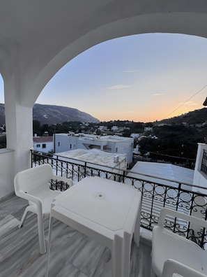 Varanda com cadeiras e mesa de plástico branco, com vista para uma área residencial com casas e montanha.