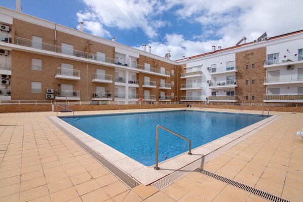 Una piscina con barandilla de metal, rodeada por una terraza con baldosas y contigua a un edificio de varios pisos con balcones.