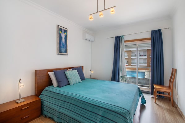 Une chambre à coucher comprenant un lit, une table de chevet, une chaise, une fenêtre avec des rideaux et une vue sur un bâtiment à l’extérieur.