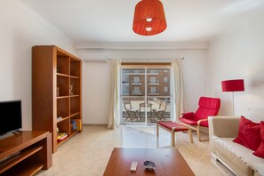 Un salon avec un téléviseur, une bibliothèque en bois, un fauteuil rouge, un canapé et une lampe rouge.