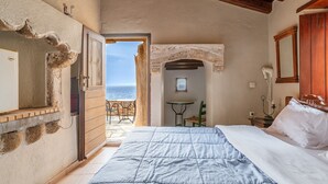 Ein Schlafzimmer mit einem Bett, einem kleinen Tisch, einem Sessel, einem Fenster mit Meerblick und einer Holzbalkendecke.
