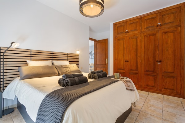 A bedroom with a bed, bedside lamps, a wooden wardrobe, and a view of a hallway.