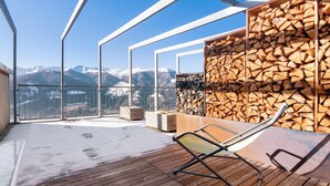 Une terrasse en bois avec une chaise, une pile de bois de chauffage et une vue sur des montagnes enneigées.