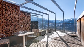 Une terrasse en bois avec une table et des chaises, un mur en pierre et une vue sur des montagnes enneigées.