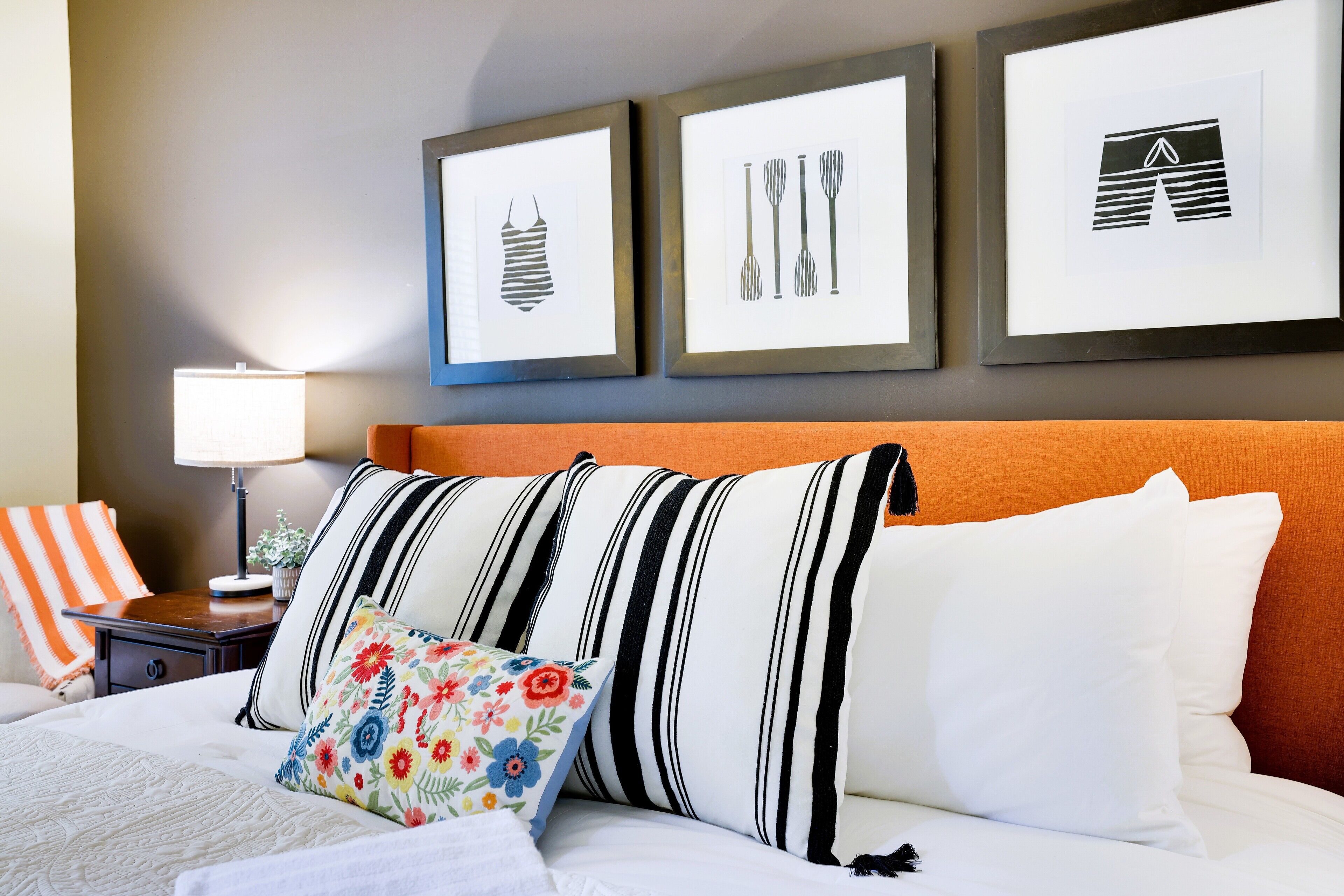 A hotel room with a bed, striped pillows, a colorful floral pillow, a nightstand with a lamp, and framed artwork on the wall.