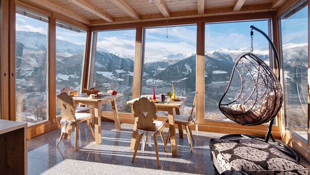 Ein Zimmer mit großen Fenstern, einem Hängesessel, einem Tisch mit Stühlen und Blick auf schneebedeckte Berge.