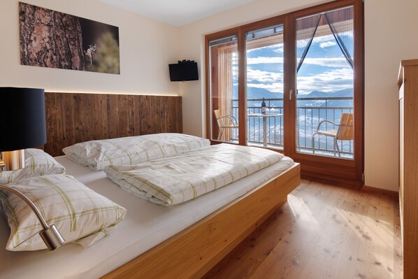 A bedroom with a bed, a wooden headboard, a mounted TV, a balcony with a view of mountains, and a chair.