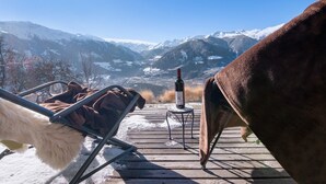 A wooden deck with a chair and a table, a bottle of wine, and a view of snow-covered mountains.