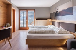 Un dormitorio con cama, escritorio, silla, radiador y vista a montañas a través de un ventanal grande.