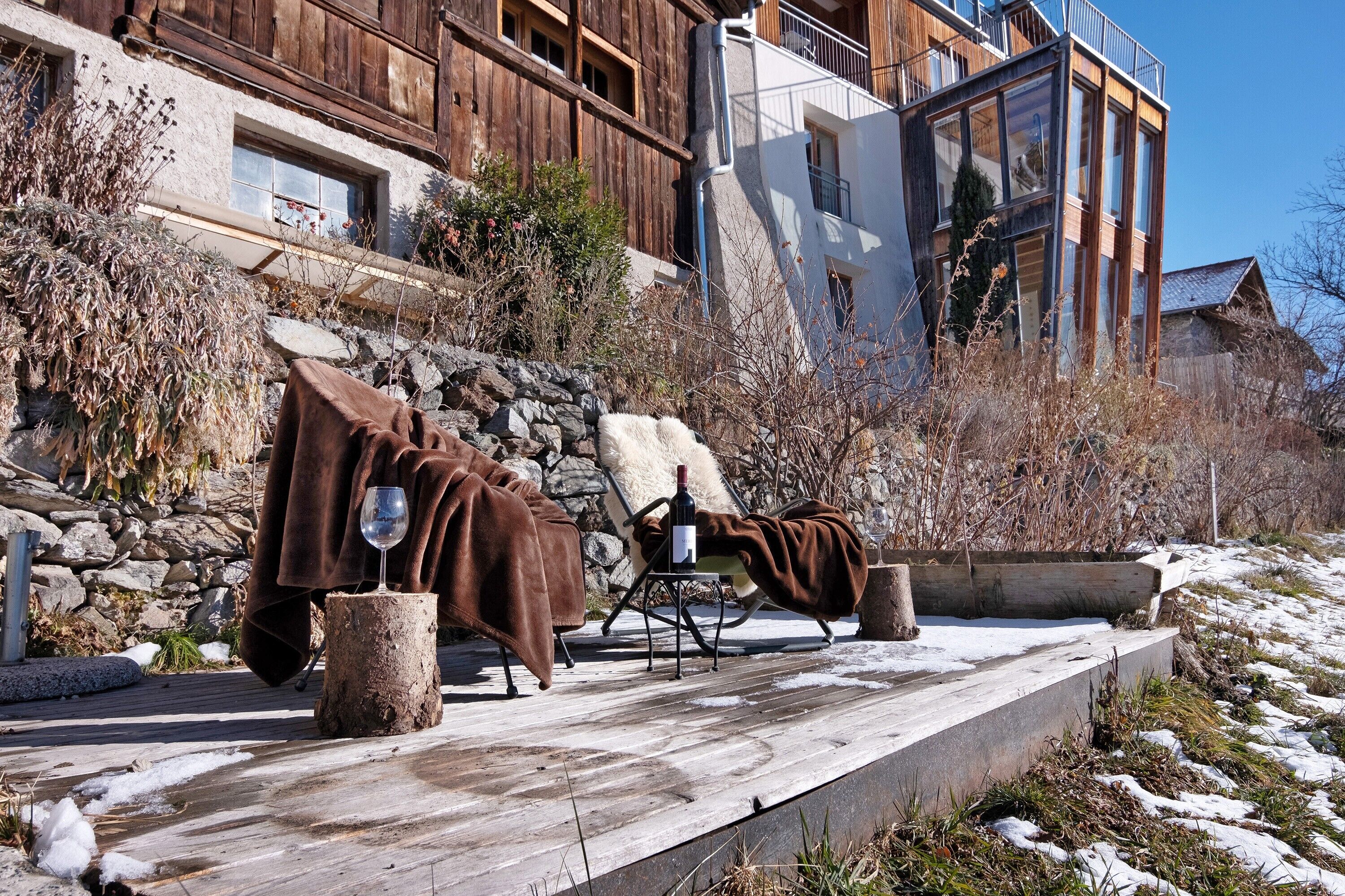 Une terrasse en bois avec deux chaises longues recouvertes de couvertures marron, une bouteille de vin et un verre.