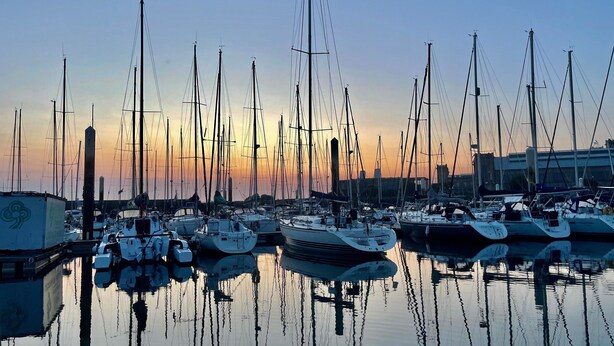 Un puerto deportivo con varias embarcaciones amarradas, agua tranquila que refleja las barcas, y un cielo despejado.