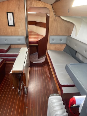 A compact interior with a wooden floor, a bench, a table, and a small window. - Sailing Boat 'Miraflores' with Sea View, Private Terrace and Wi-Fi (Cherbourg)