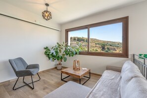 Una habitación con vistas a un paisaje montañoso, una silla gris, un sofá blanco, una mesita de madera y una planta en maceta.