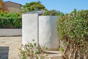 Une structure en béton surmontée d’un toit métallique, entourée de verdure et bordée d’une zone dallée en pierre.