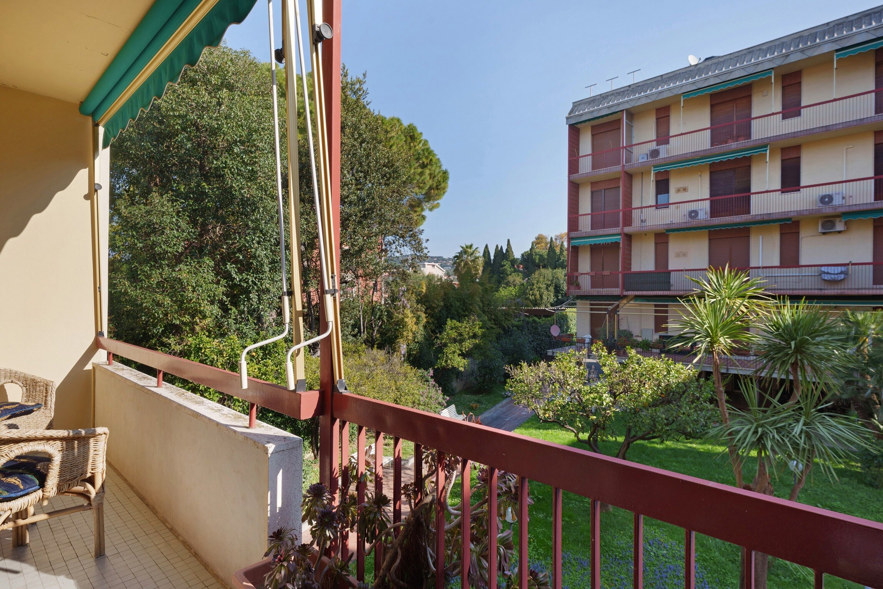 Ein Balkon mit Blick auf ein mehrstöckiges Gebäude, Grünflächen und einen klaren Himmel.