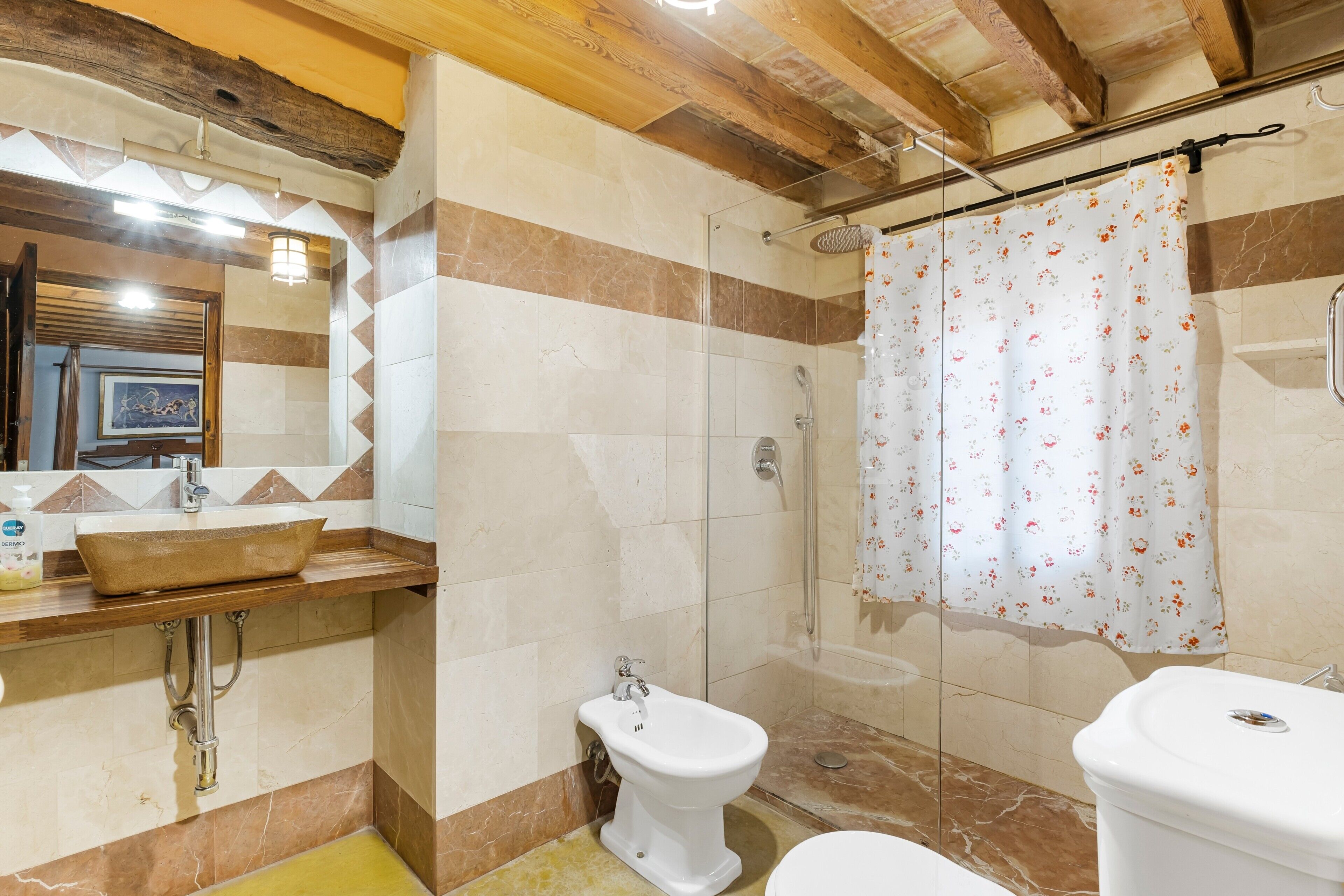 Un baño con tocador de madera, lavamanos sobre encimera, bidé, ducha con cortina estampada con motivos florales y inodoro.