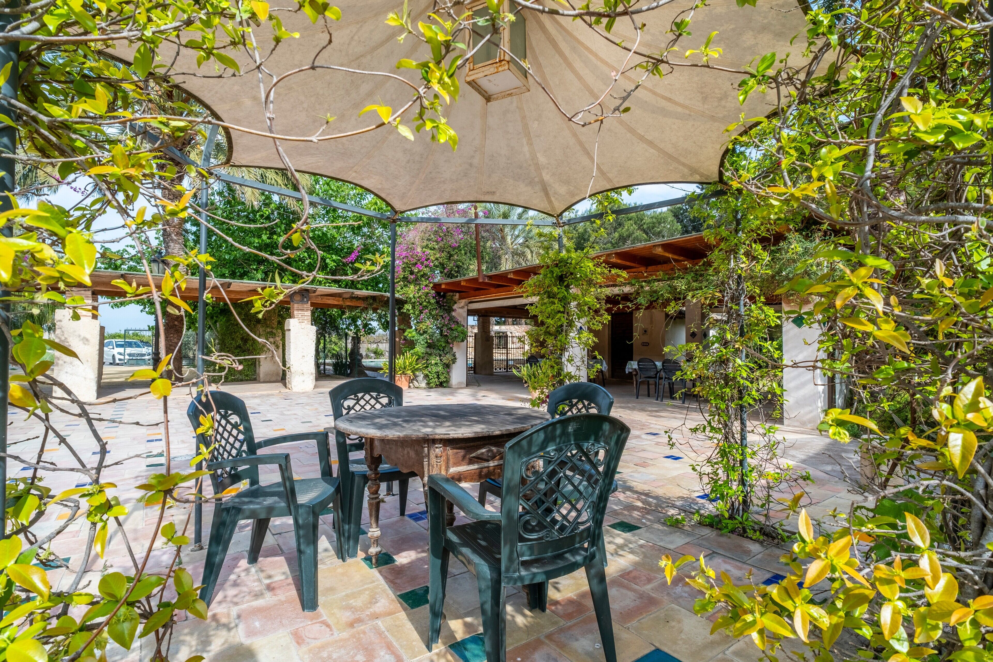 Un patio al aire libre con una mesa redonda y sillas, rodeado de follaje y cubierto por un toldo grande.