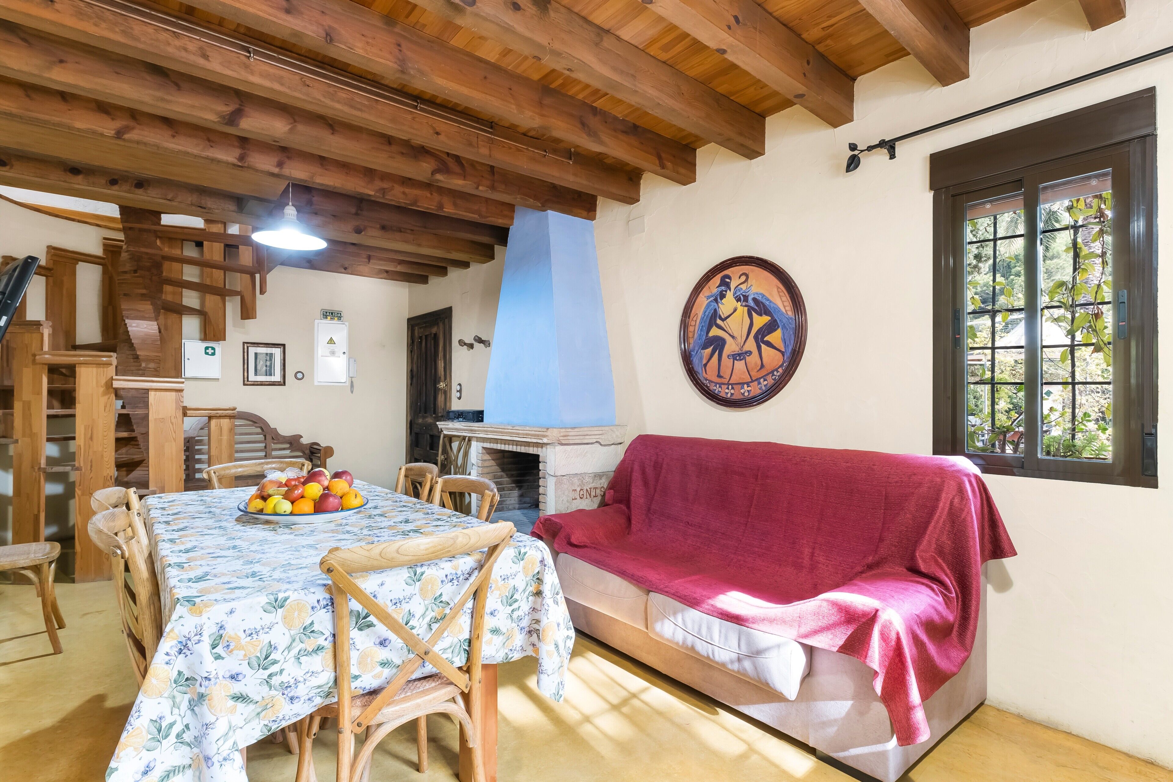 Un coin repas avec une table dressée pour quatre personnes, un canapé recouvert d’un plaid rouge, une cheminée et un escalier en bois.