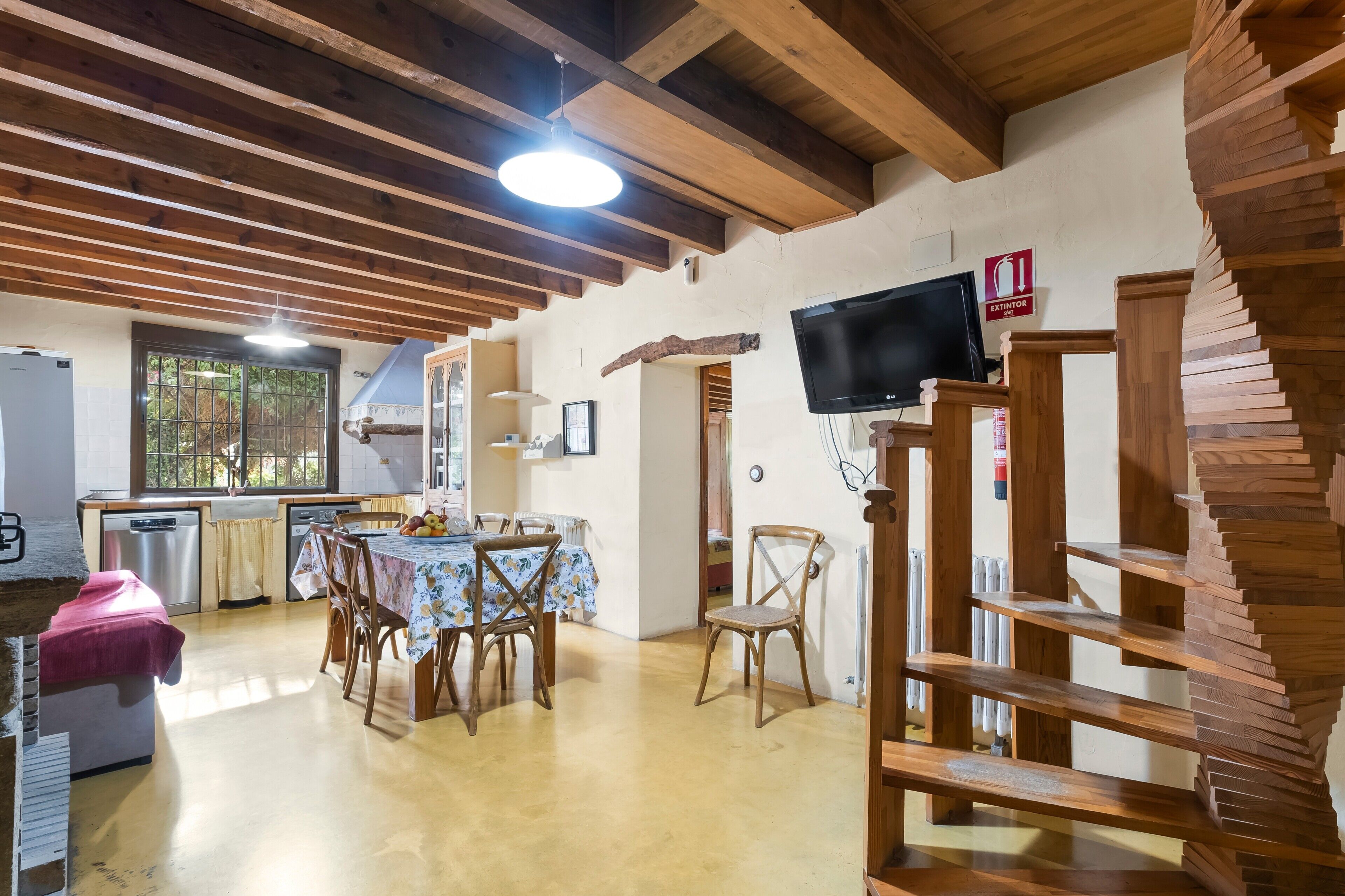 Un interior rústico con vigas de madera a la vista, una zona de comedor con mesa y sillas, una cocina con lavavajillas y un televisor montado en la pared.