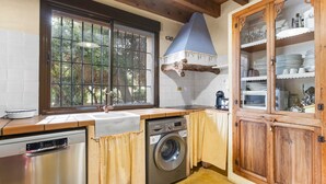 A kitchen with a washing machine, a sink, a window with a view of trees, and a wooden cabinet with dishes. - Country House 'La Carrasca - Limón' with Shared Pool, Wi-Fi and Air Conditioning (Catral)
