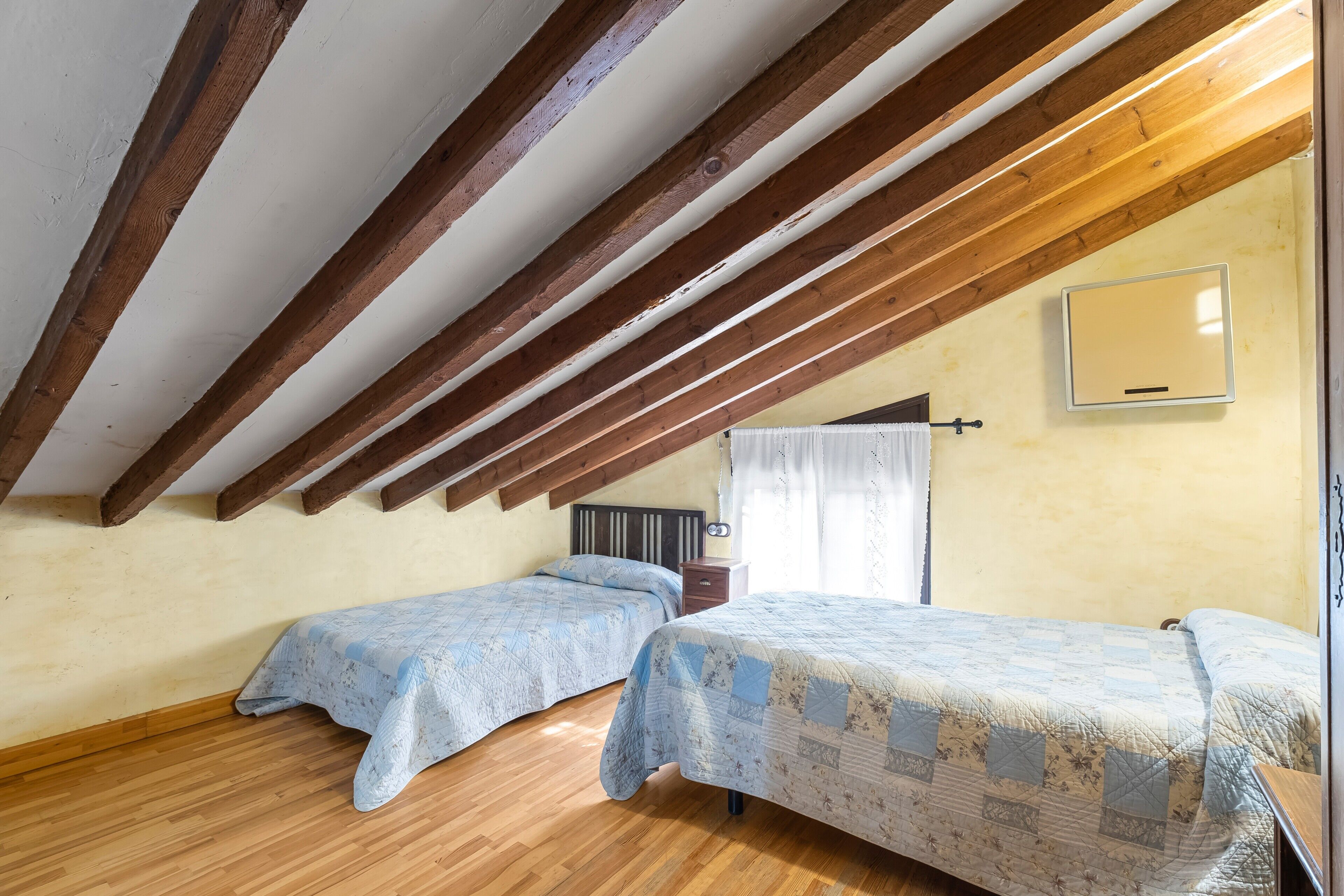 A room with wooden beams, two beds with patterned quilts, a bedside table, and a mirror on the wall.