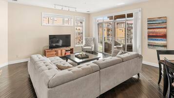 A living room with a sectional sofa, a flat-screen TV, a coffee table, and a balcony with a view of buildings.